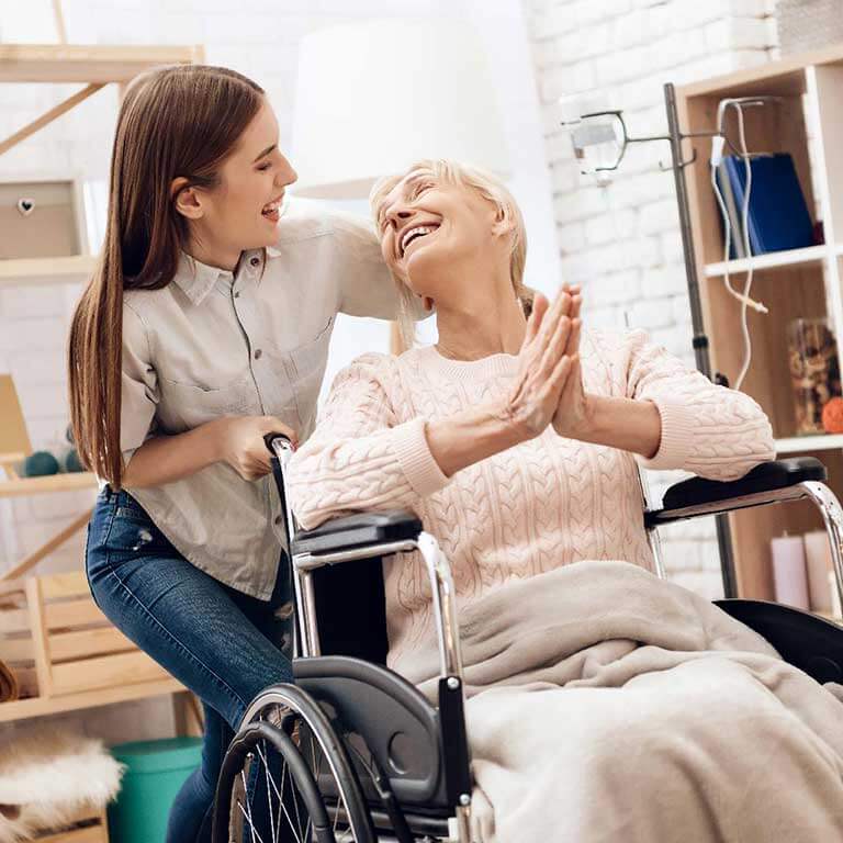 Happy senior woman in a wheelchair high-fiving her caregiver in a cozy home setting, symbolizing compassionate in-home care services for the elderly.